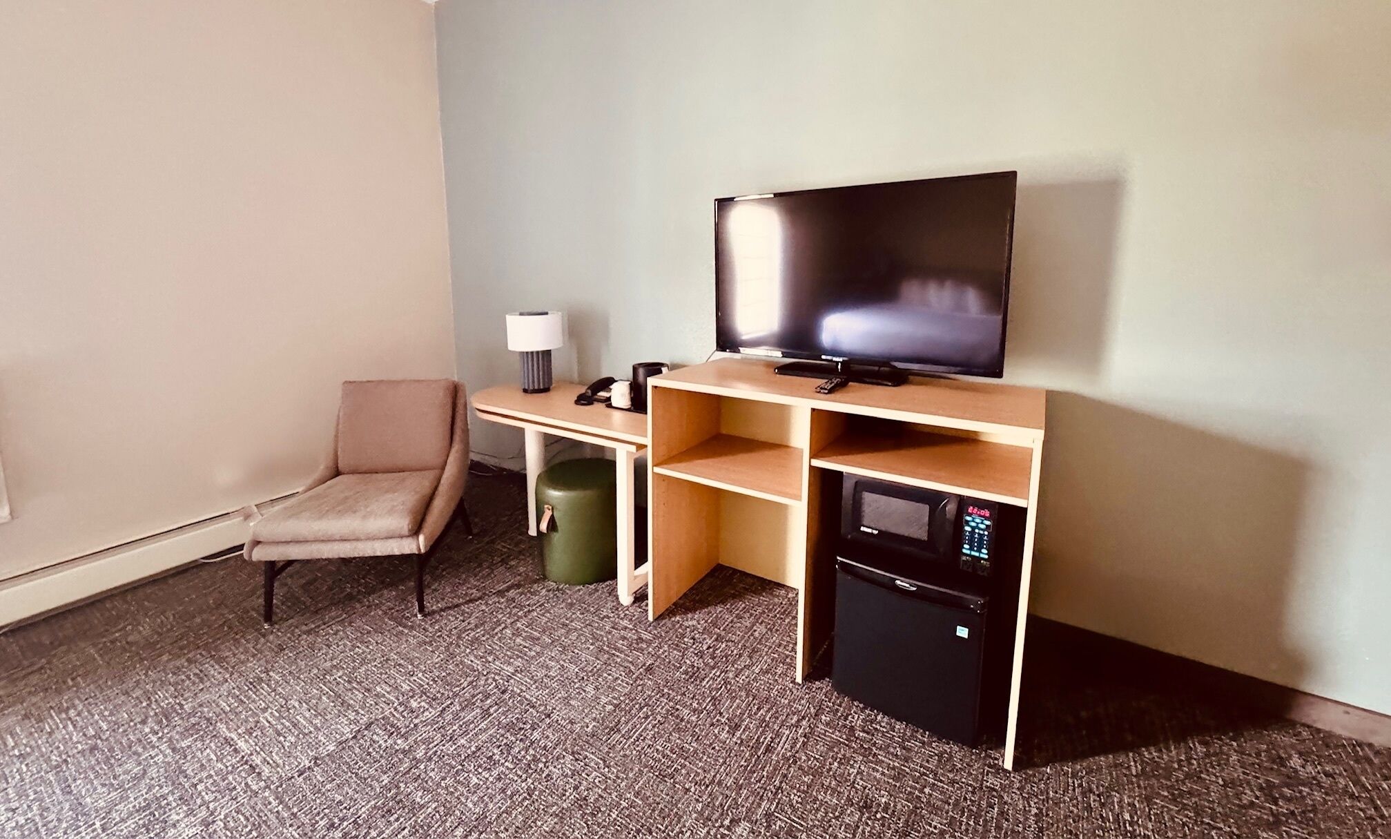 A hotel room with a single bed, a desk with a flat-screen TV, a chair, a trash bin, and a mini-fridge.