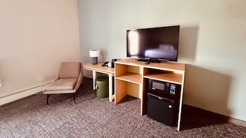 Habitación de hotel con una cama individual, un escritorio con televisor de pantalla plana, una silla, un cesto de basura y un mini refrigerador.