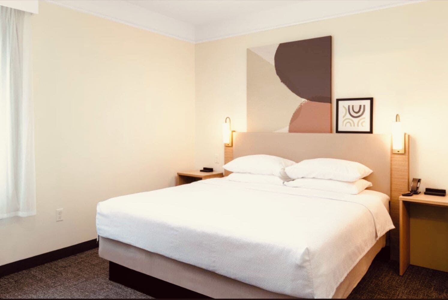 A hotel room with a large bed, bedside tables, a wall-mounted artwork, and a window with curtains.