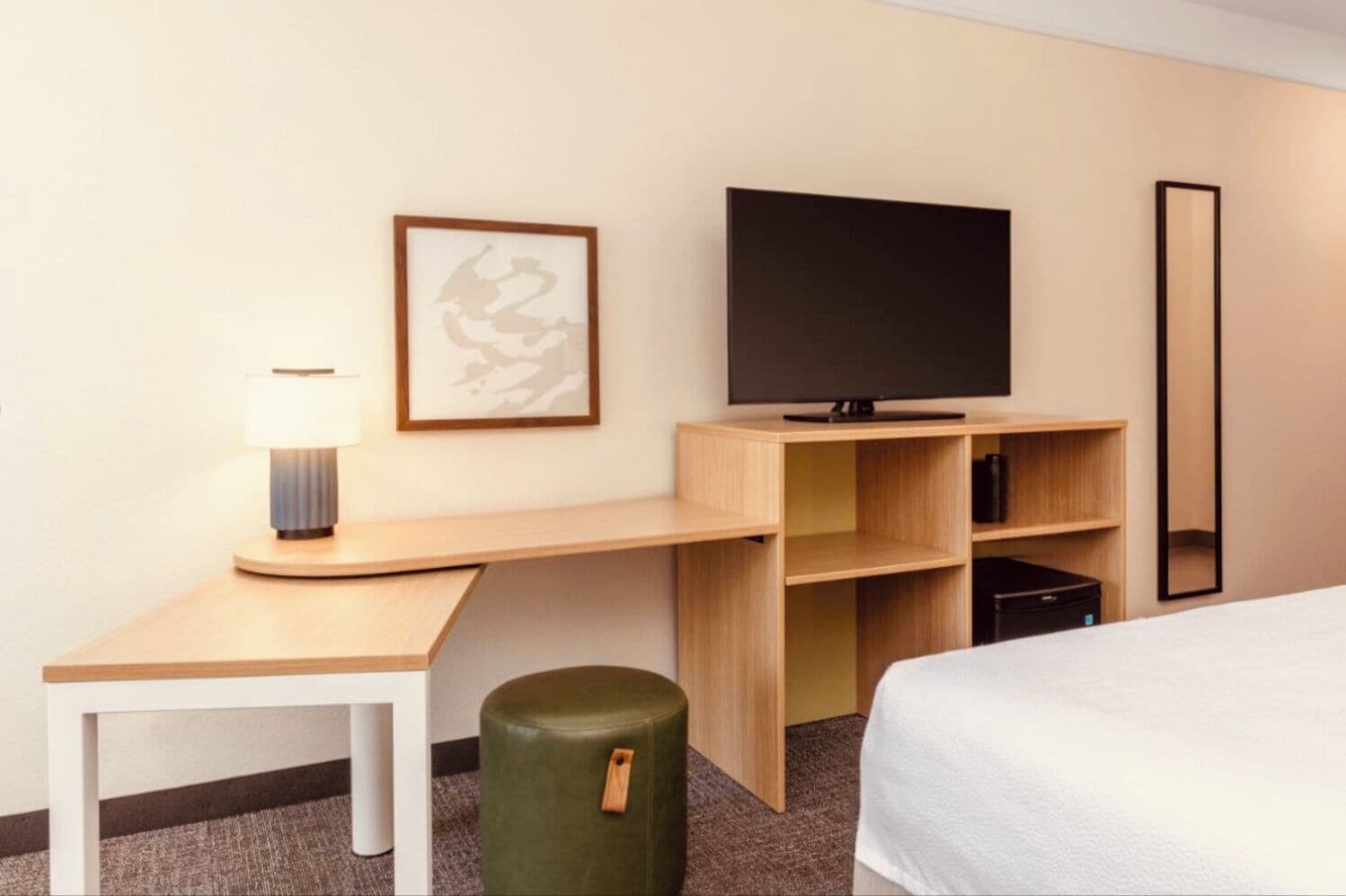 A hotel room with a bed, a desk with a lamp, a television, a mirror, and a green ottoman.