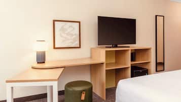 Une chambre d’hôtel avec un lit, un bureau doté d’une lampe, une télévision, un miroir et un pouf vert.