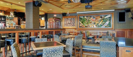 A restaurant with wooden tables and patterned chairs, a bar area, and a large framed painting on the wall.