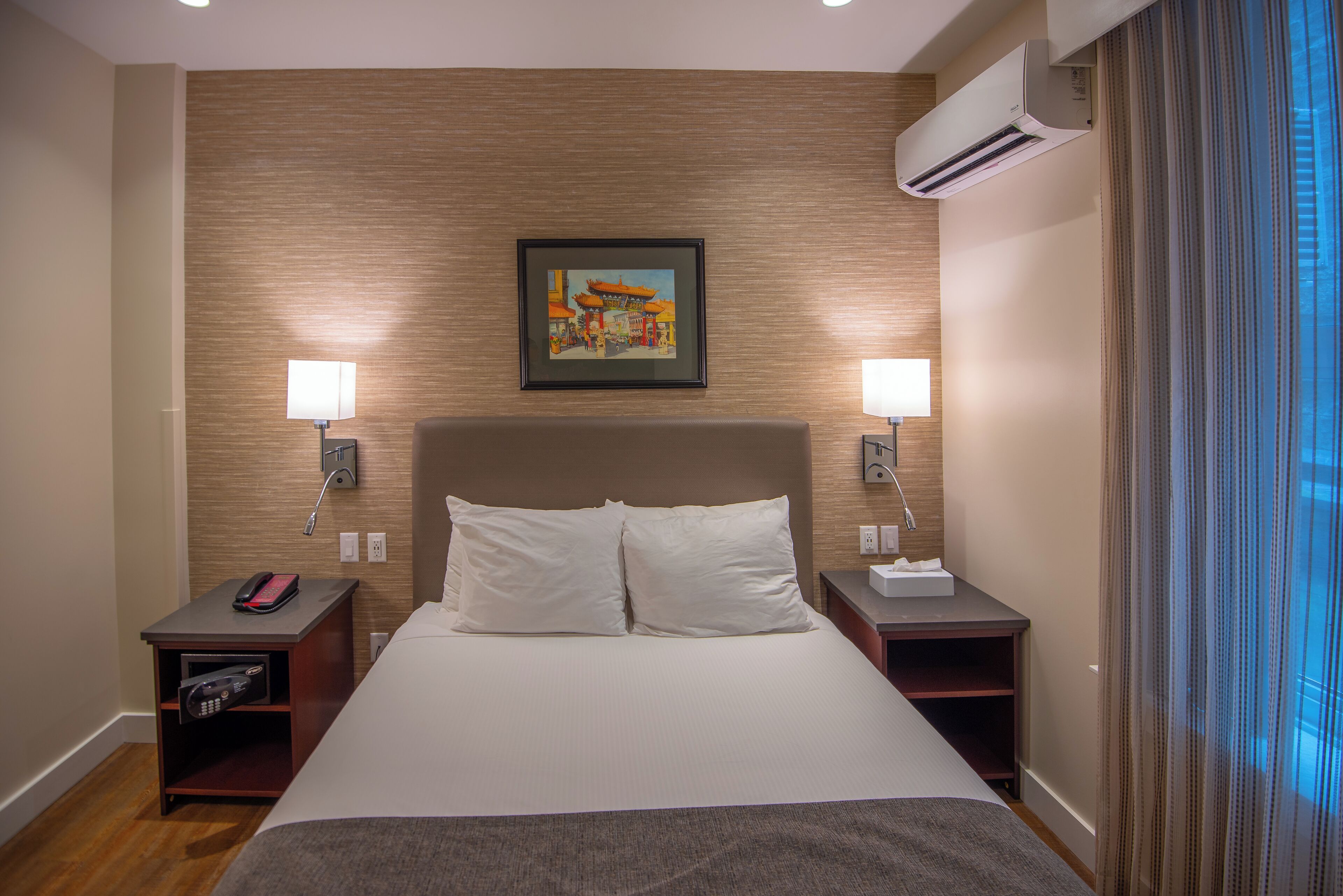 Hotelkamer met een groot bed, twee nachtkastjes, een wandairconditioner en een schilderij boven het bed.