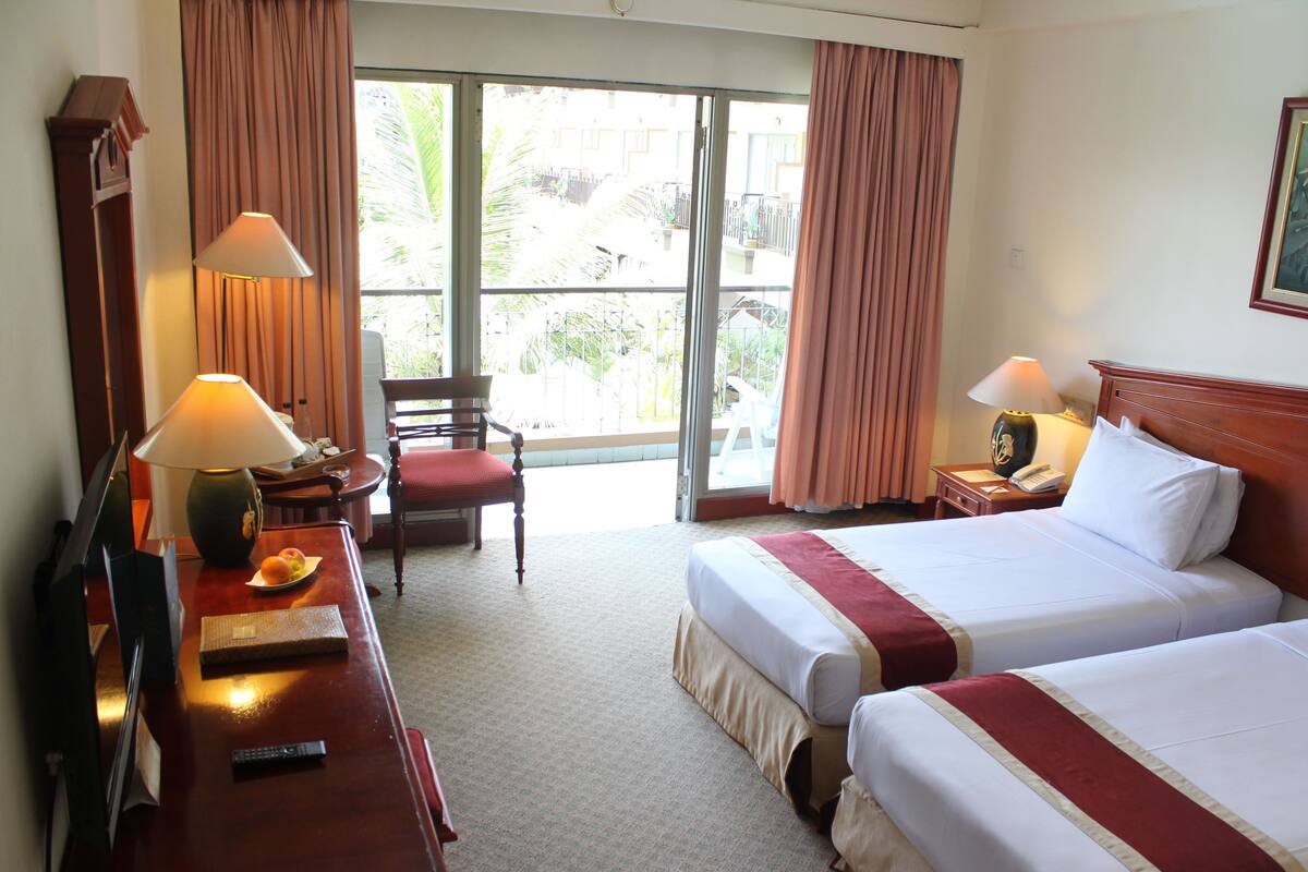 A hotel room with two beds, a desk with a lamp, a chair, and a large window with curtains.