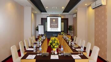 Un salón de conferencias con una mesa larga, sillas blancas y una pantalla proyectora mostrando 'The Jayakarta Suites Bandung'.