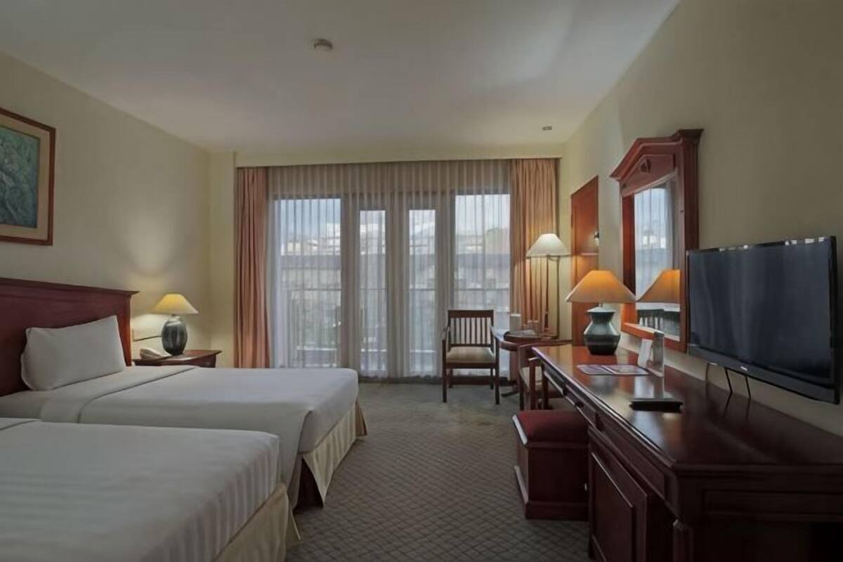 A hotel room with two beds, a desk with a chair, a TV, and a large window with curtains.