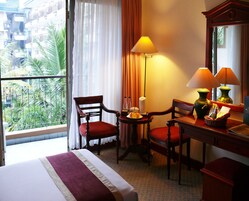 Une chambre d’hôtel comprenant un lit, deux chaises, un bureau avec une lampe, un miroir et une vue sur un balcon agrémenté de plantes.