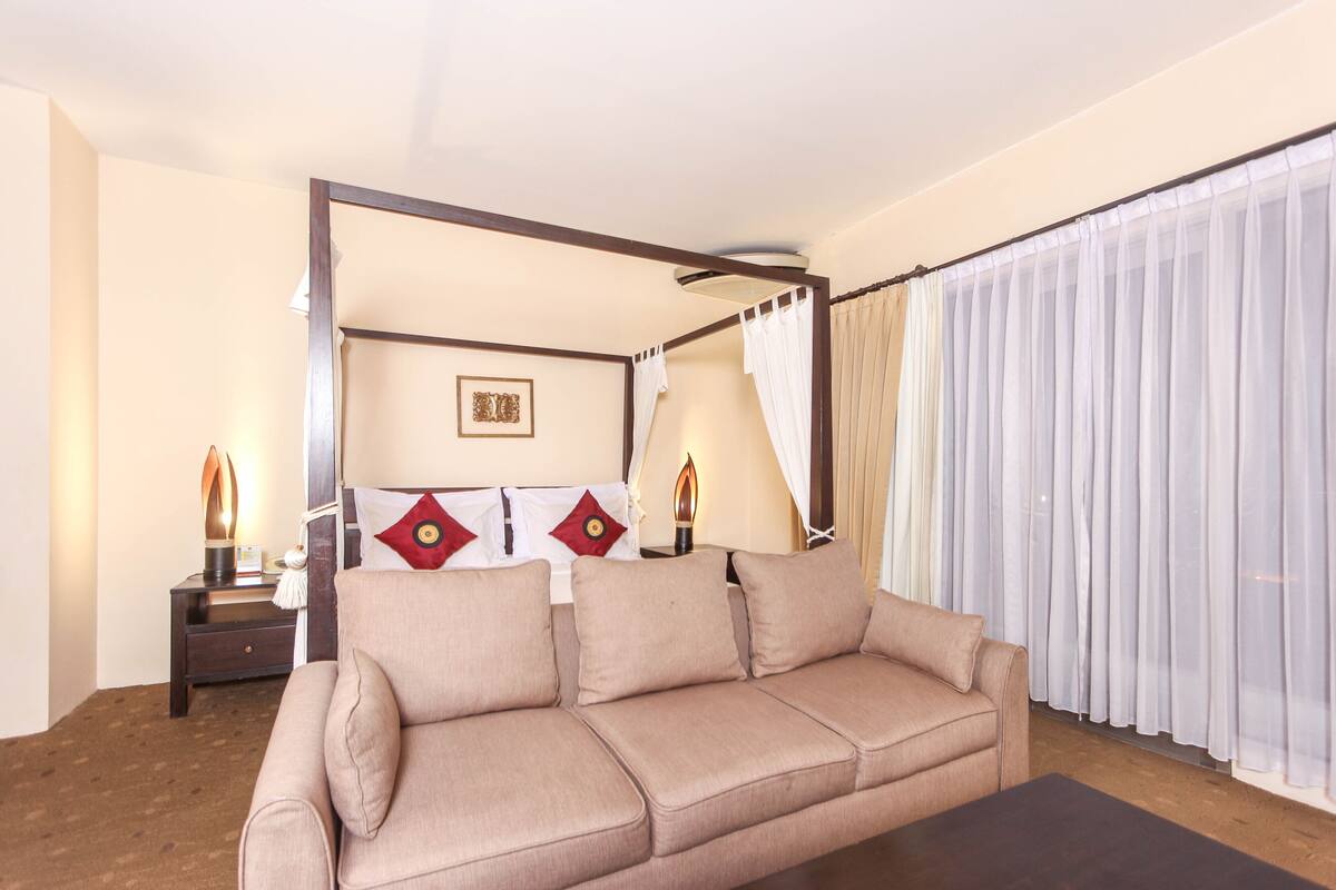 A hotel room with a four-poster bed, a sofa, a nightstand with lamps, and sheer curtains.