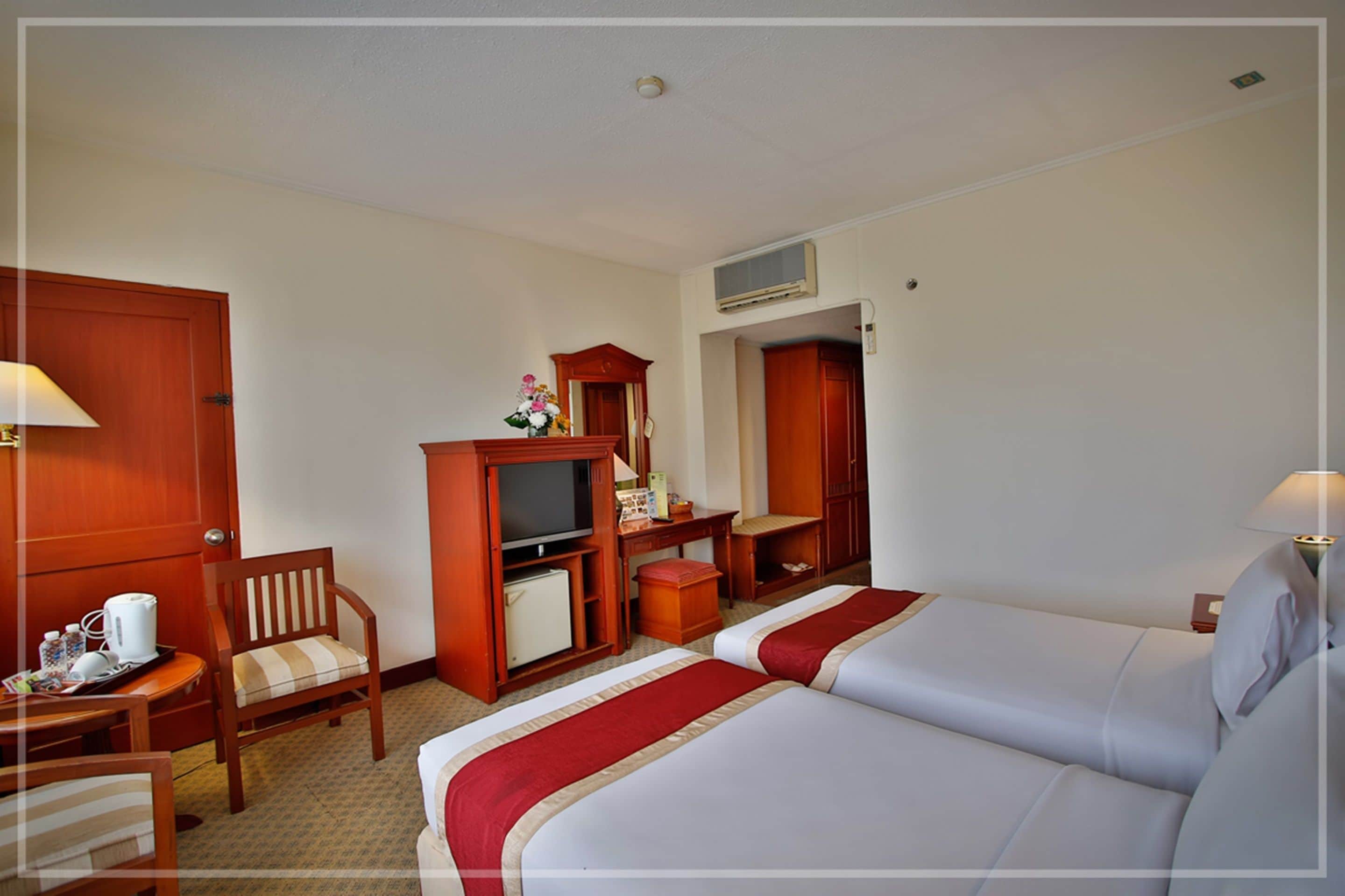 A hotel room with two beds, a wooden desk, a television, a mirror, and a small table with a lamp.