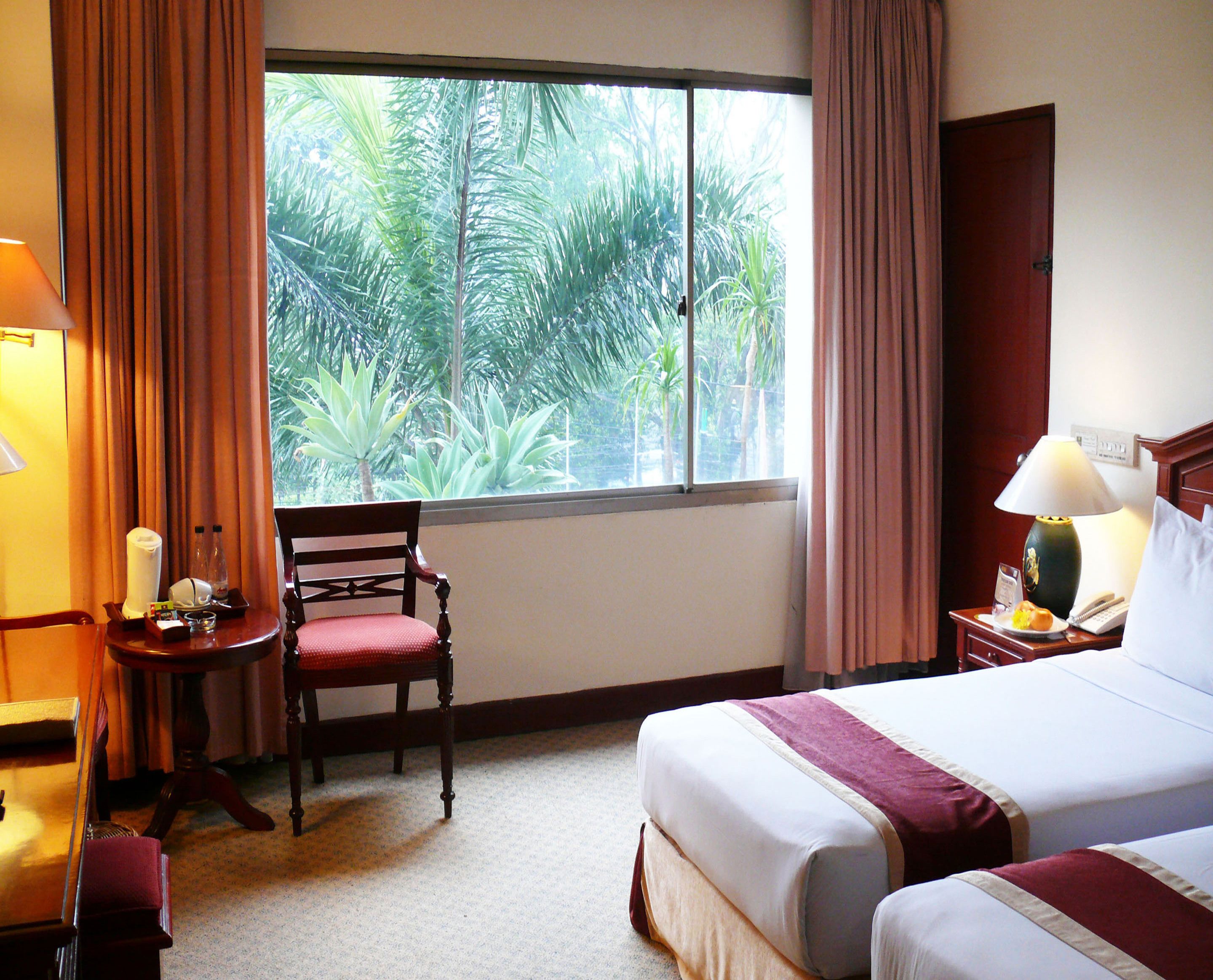 Habitación de hotel con cama, una silla, una mesita, lámpara y ventana con cortinas.
