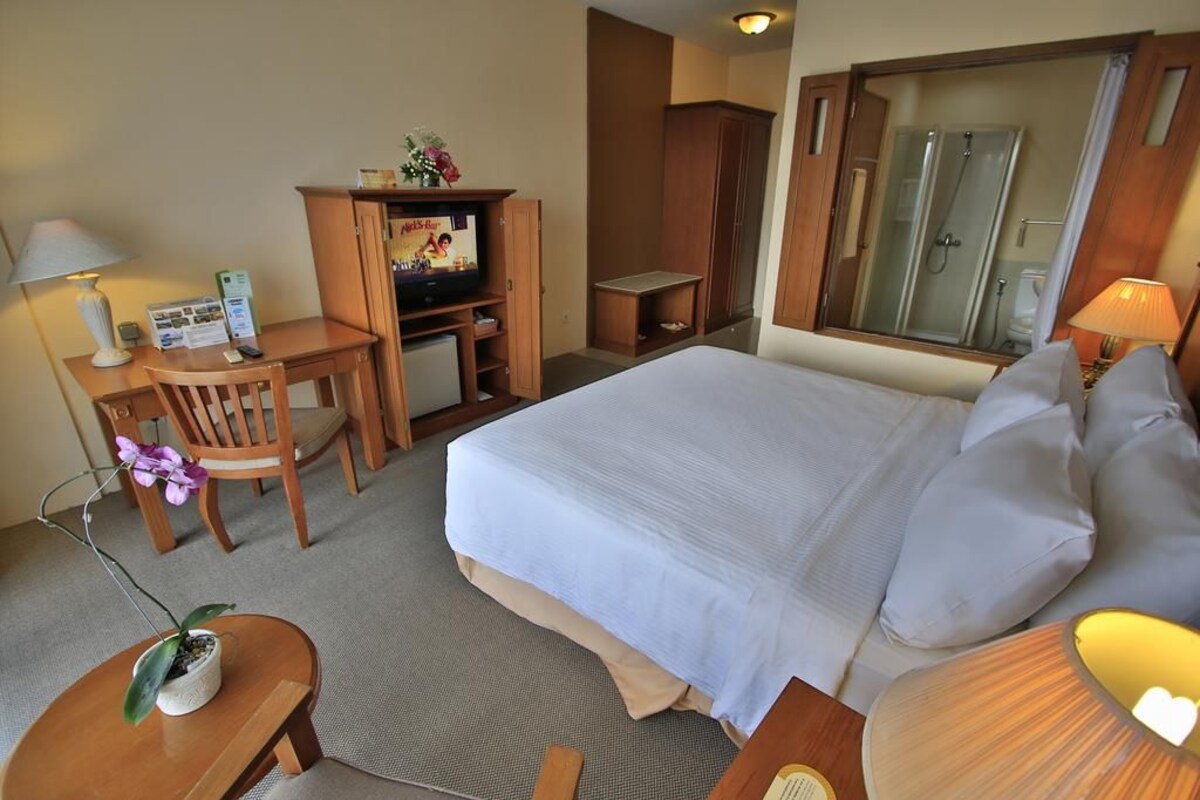 A hotel room with a bed, a desk with a television, a chair, a small table with a lamp, and a shower area.