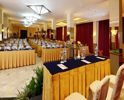 Un salón de conferencias con varias mesas dispuestas para una reunión, equipado con botellas de agua, vasos y blocs de notas.