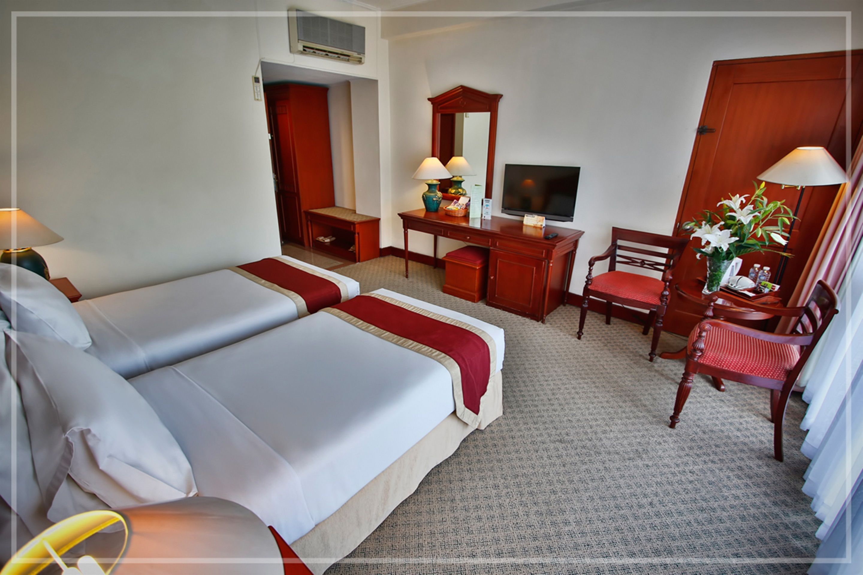A hotel room with two beds, a desk with a TV, a chair, and a small table with a vase of flowers.