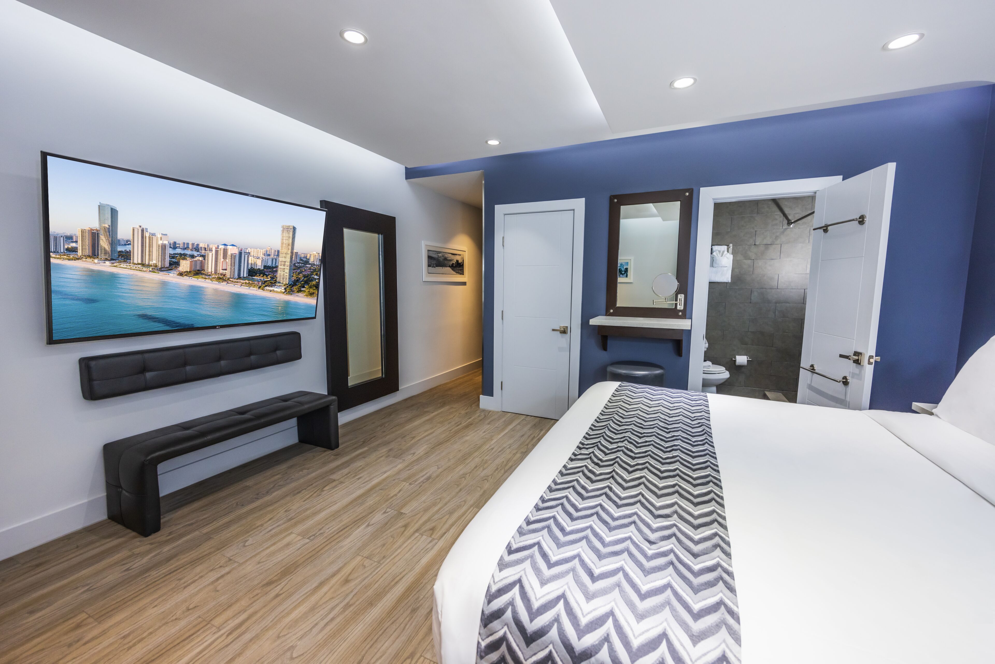 A modern hotel room with a large bed, a flat-screen TV, a bench, a bathroom with a shower, and a cityscape view.