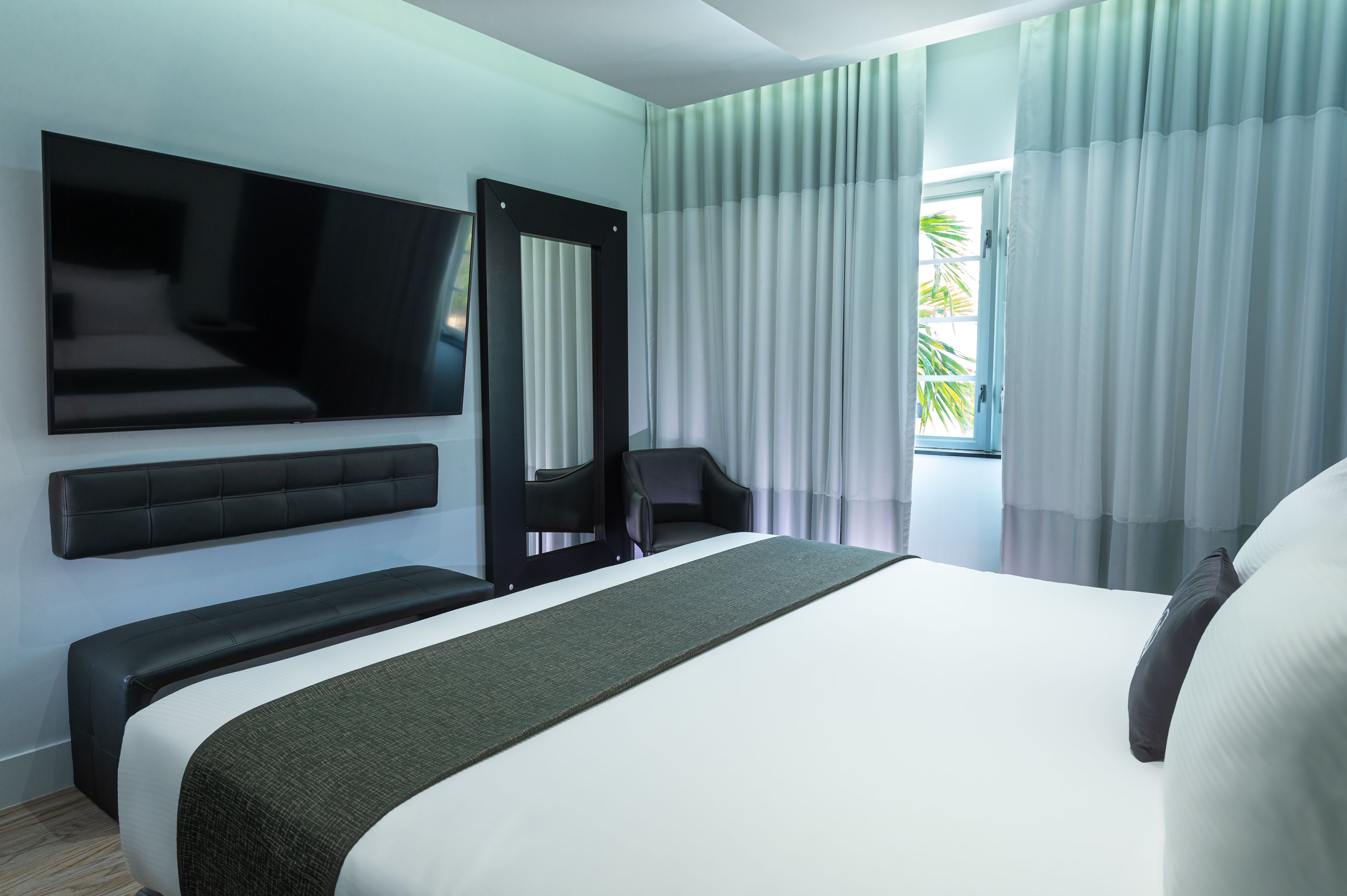 A modern hotel room with a large bed, a flat-screen TV mounted on the wall, a seating area with a bench, and a window with curtains.