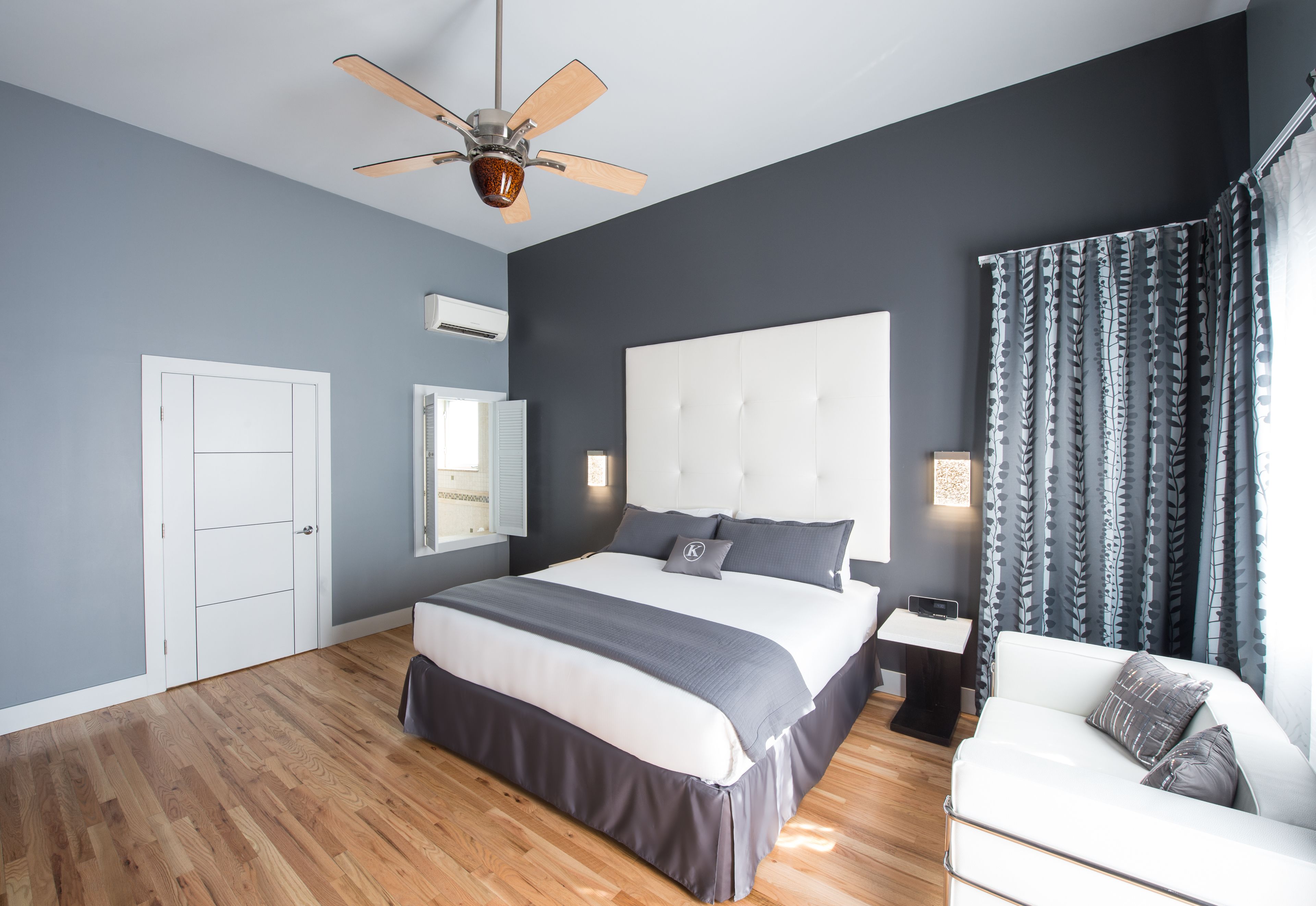 Un dormitorio moderno con una cama grande, un ventilador de techo, un cabecero blanco, una mesita de noche y un sofá blanco.