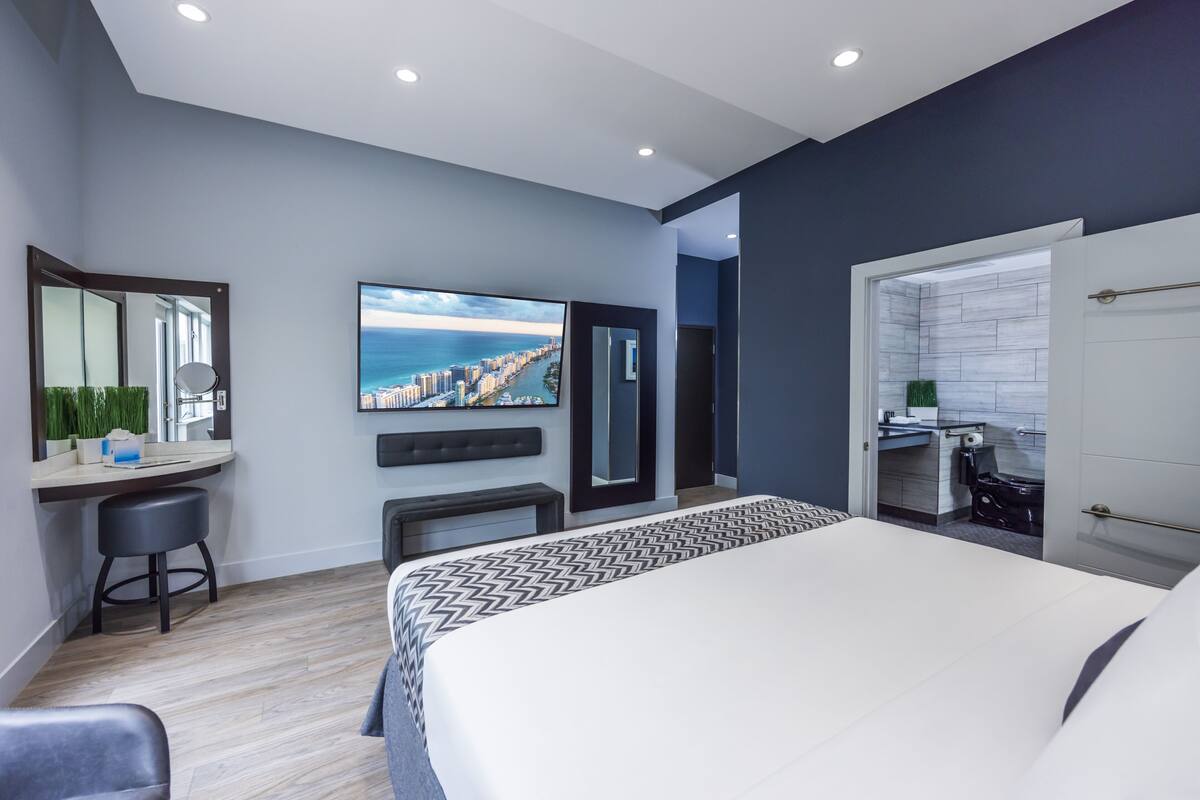 A modern bedroom with a large bed, a desk, a chair, a mirror, and a TV displaying a cityscape.