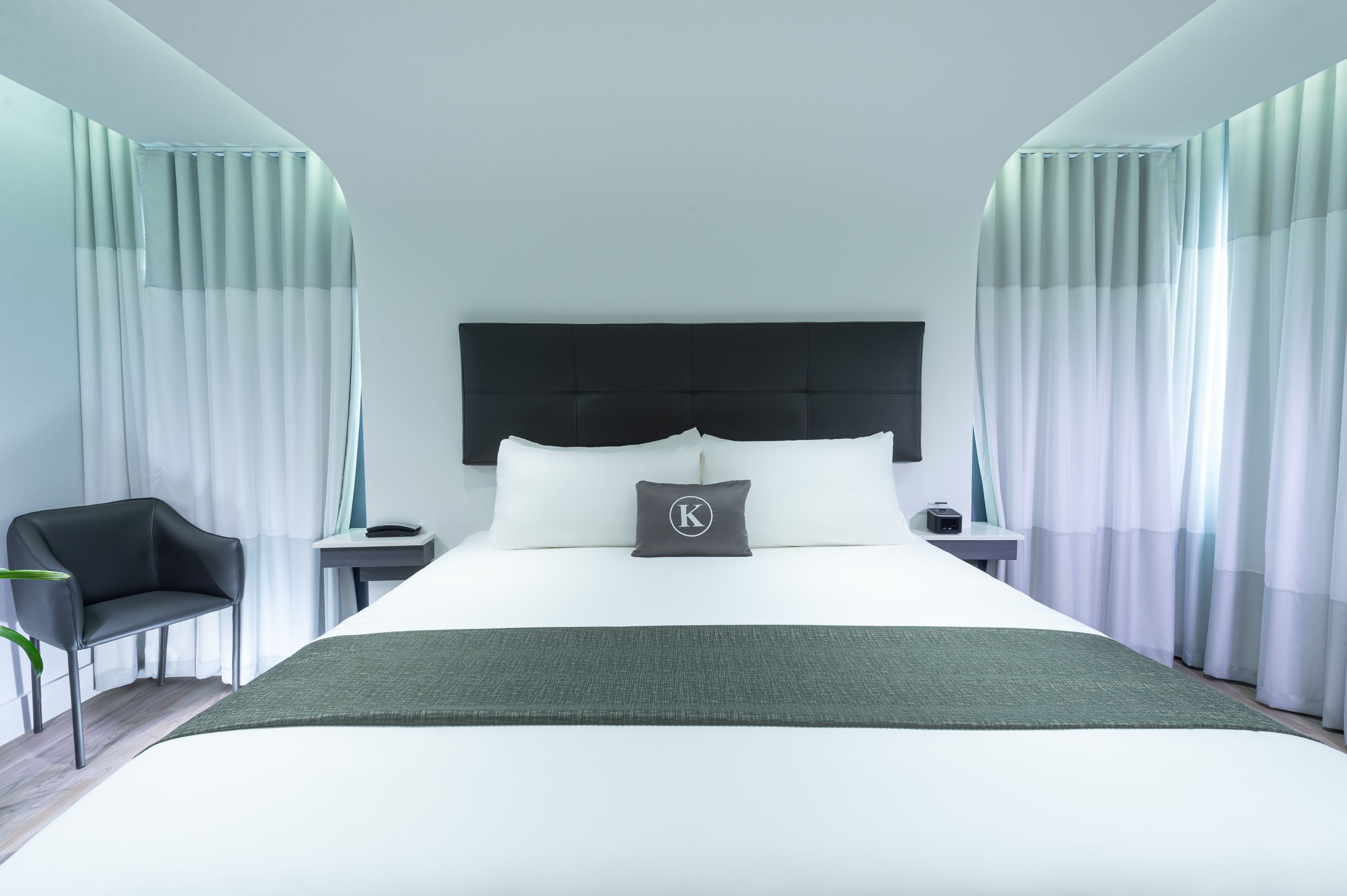 Una habitación de hotel moderna con una cama grande, un cabecero negro, una mesita de noche, una silla y cortinas de gasa.