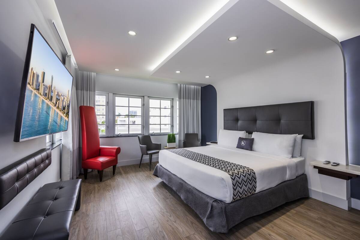 A modern hotel room with a large bed, a red chair, a desk, and a TV displaying a cityscape.