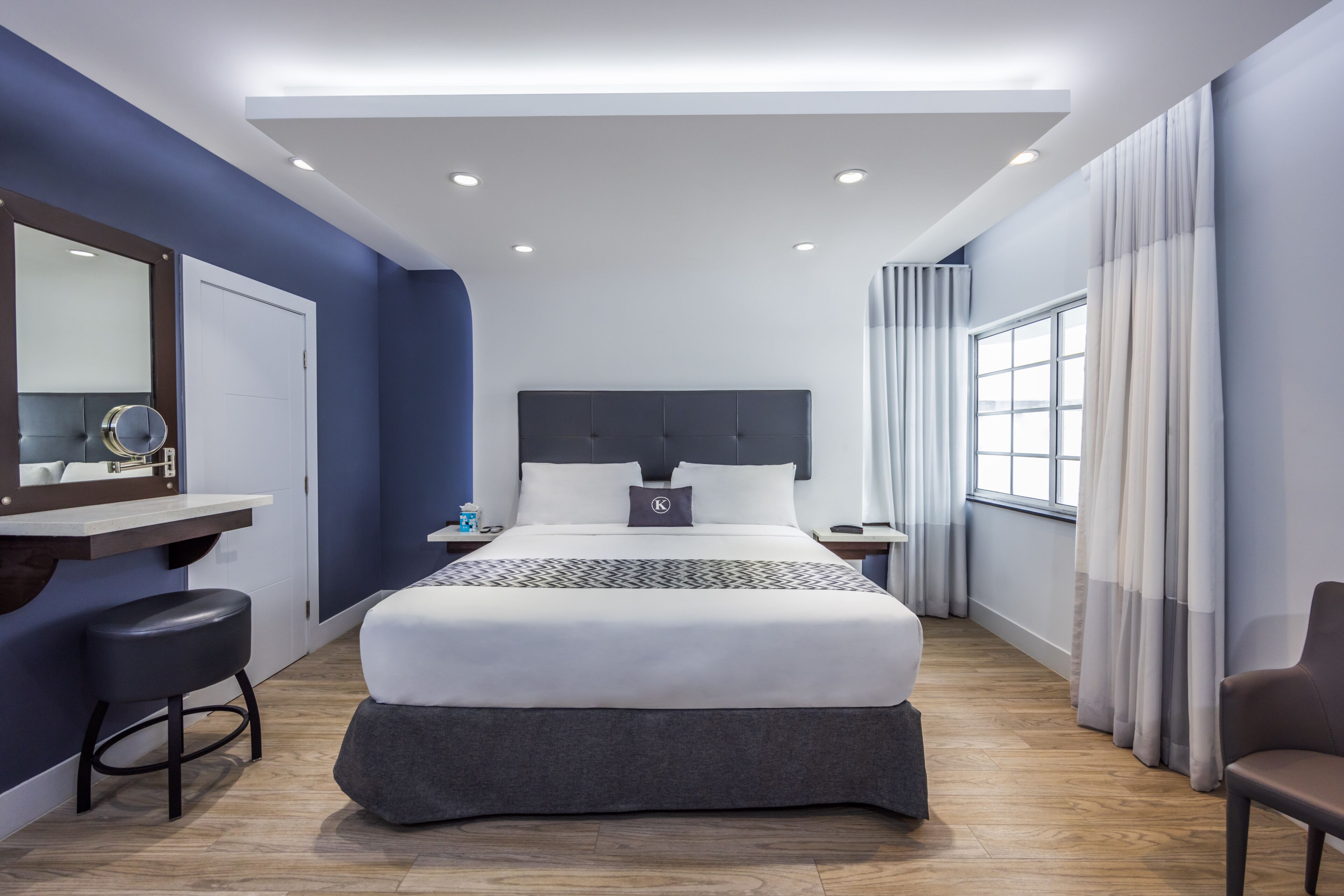 A modern hotel room with a large bed, a desk with a chair, a vanity with a mirror, and a window with curtains.