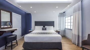 A modern hotel room with a large bed, a desk with a chair, a vanity with a mirror, and a window with curtains.