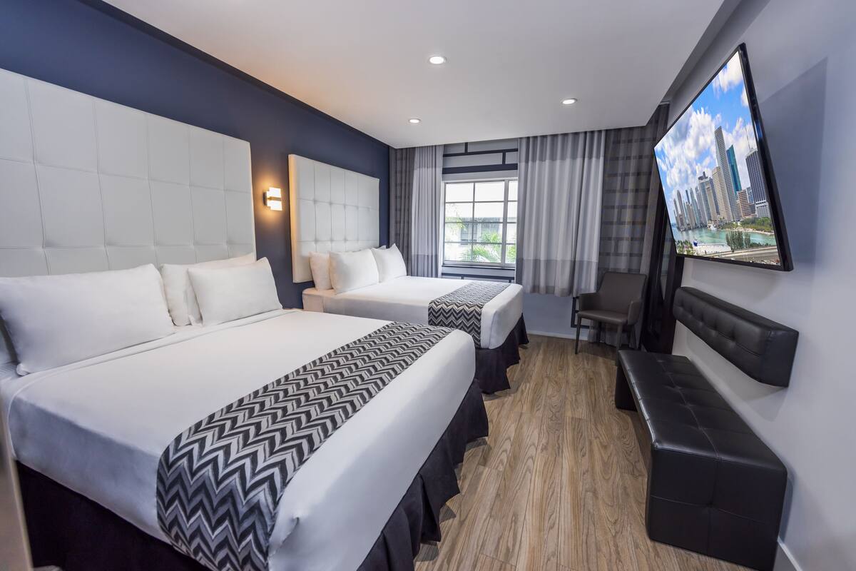 A hotel room with two beds, a TV, a bench, and a city view.