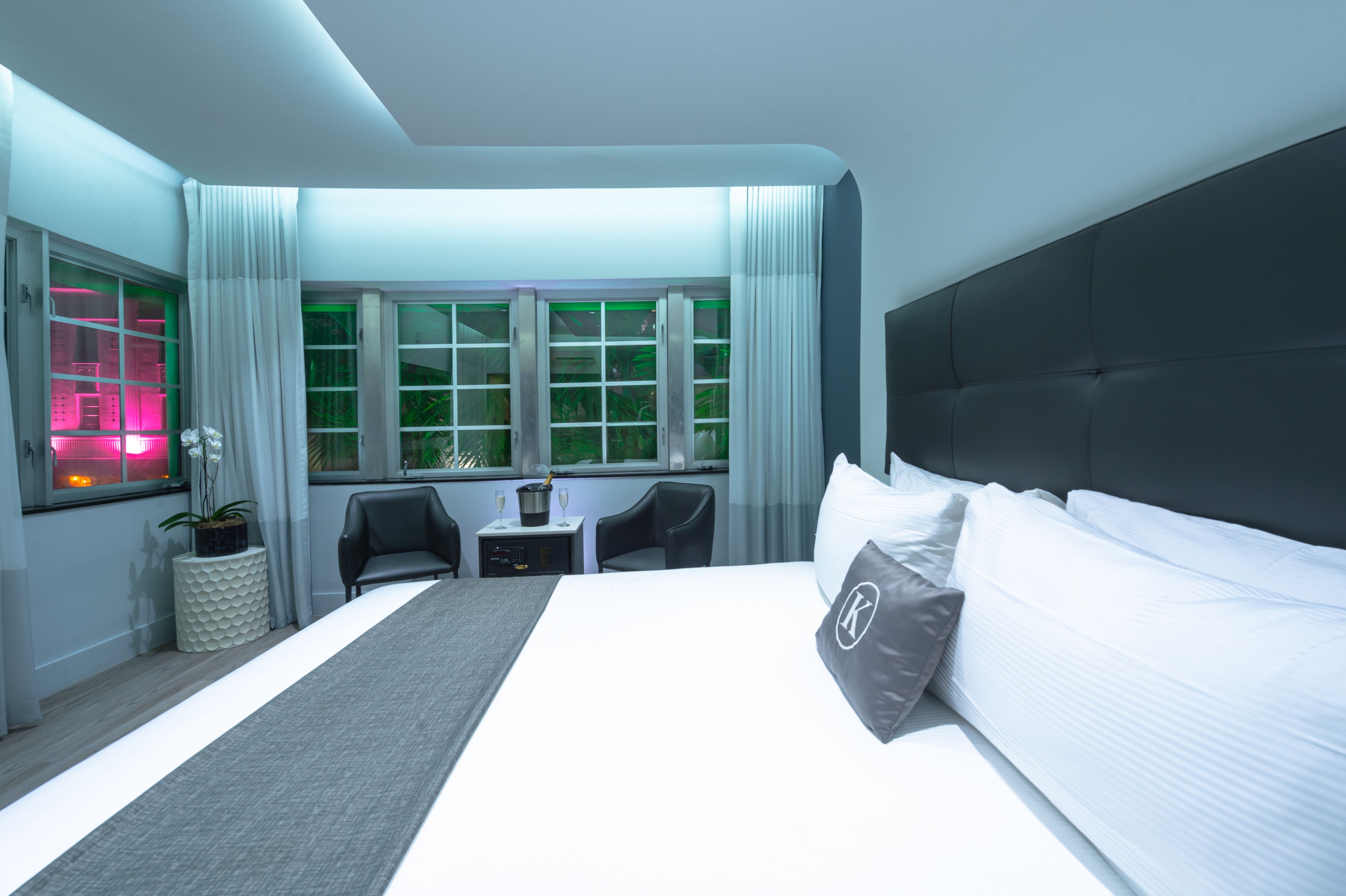Ein modernes Hotelzimmer mit einem großen Bett, einem Schreibtisch mit Stuhl, einem kleinen Tisch mit Lampe und einem Fenster mit Vorhängen.