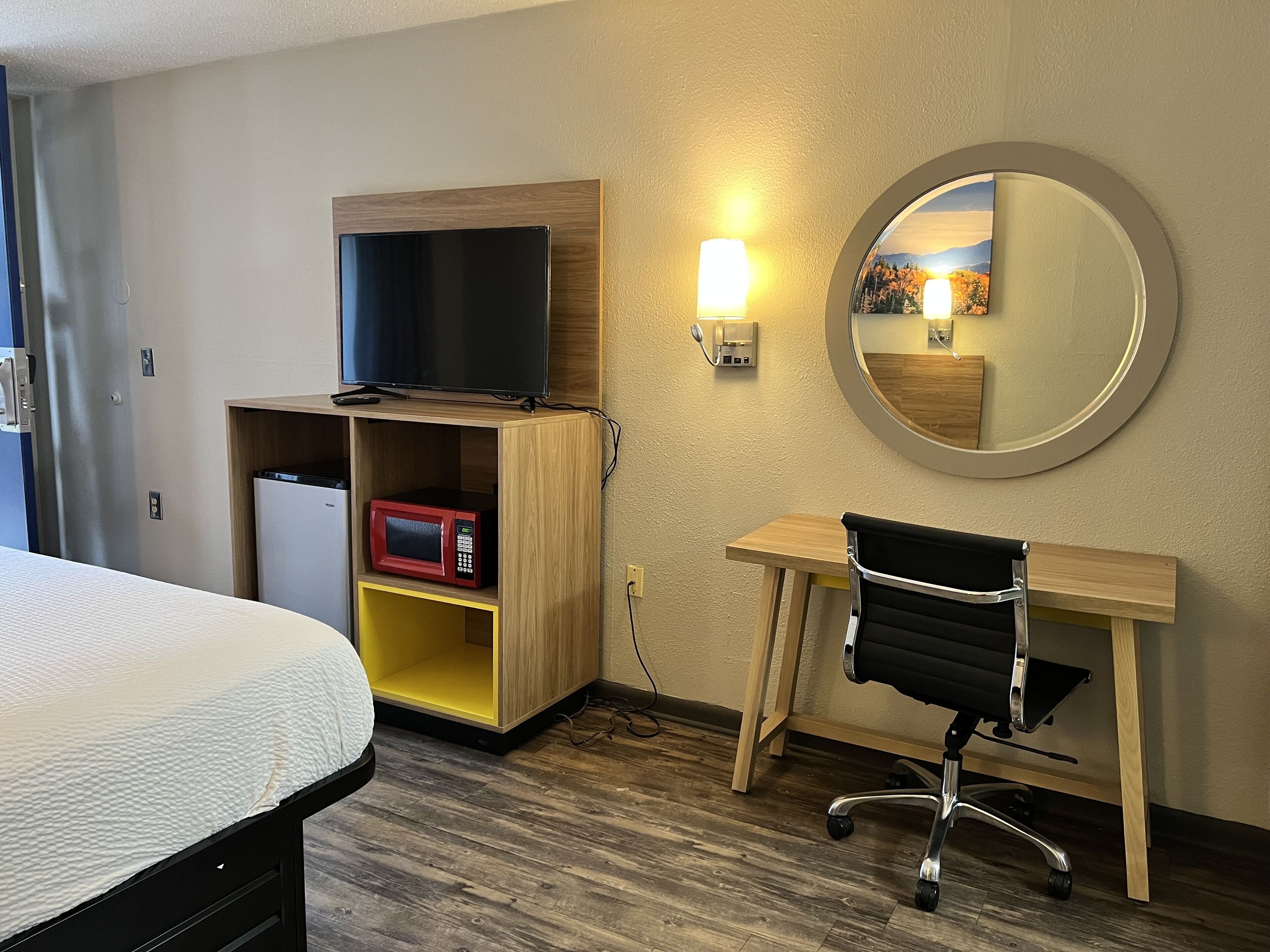 A hotel room with a bed, a desk with a chair, a TV, a microwave, and a mirror.