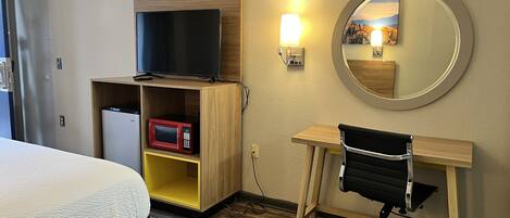 A hotel room with a bed, a desk with a chair, a TV, a microwave, and a mirror.