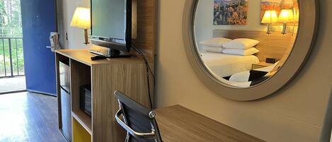 Een hotelkamer met een bureau, stoel, televisie en een ronde spiegel die een bed met kussens en een schilderij van een berglandschap weerspiegelt.
