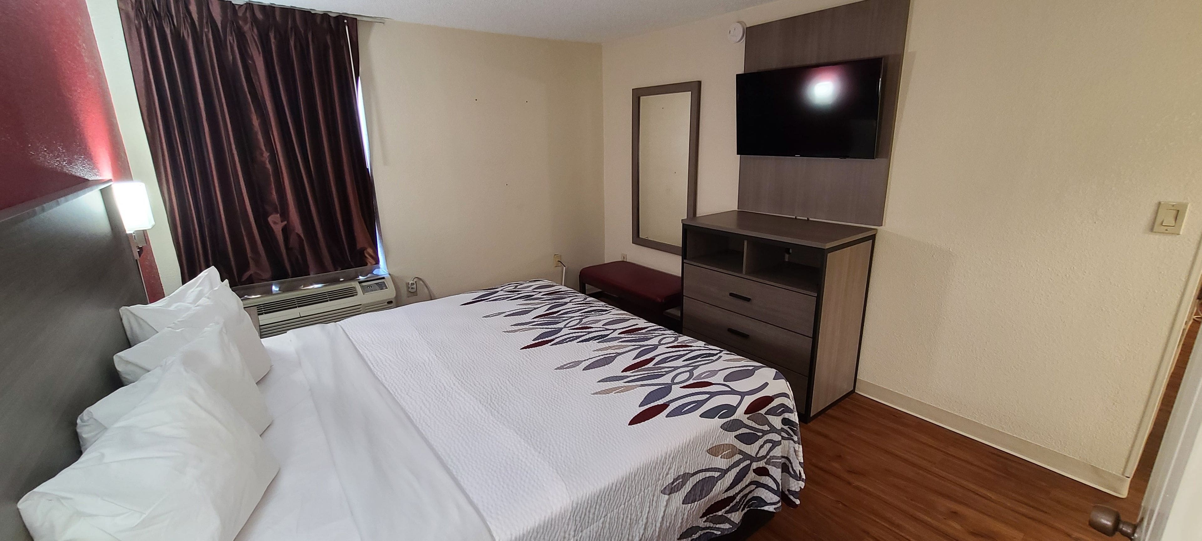 Habitación de hotel con cama, escritorio, televisor y ventana con cortinas.