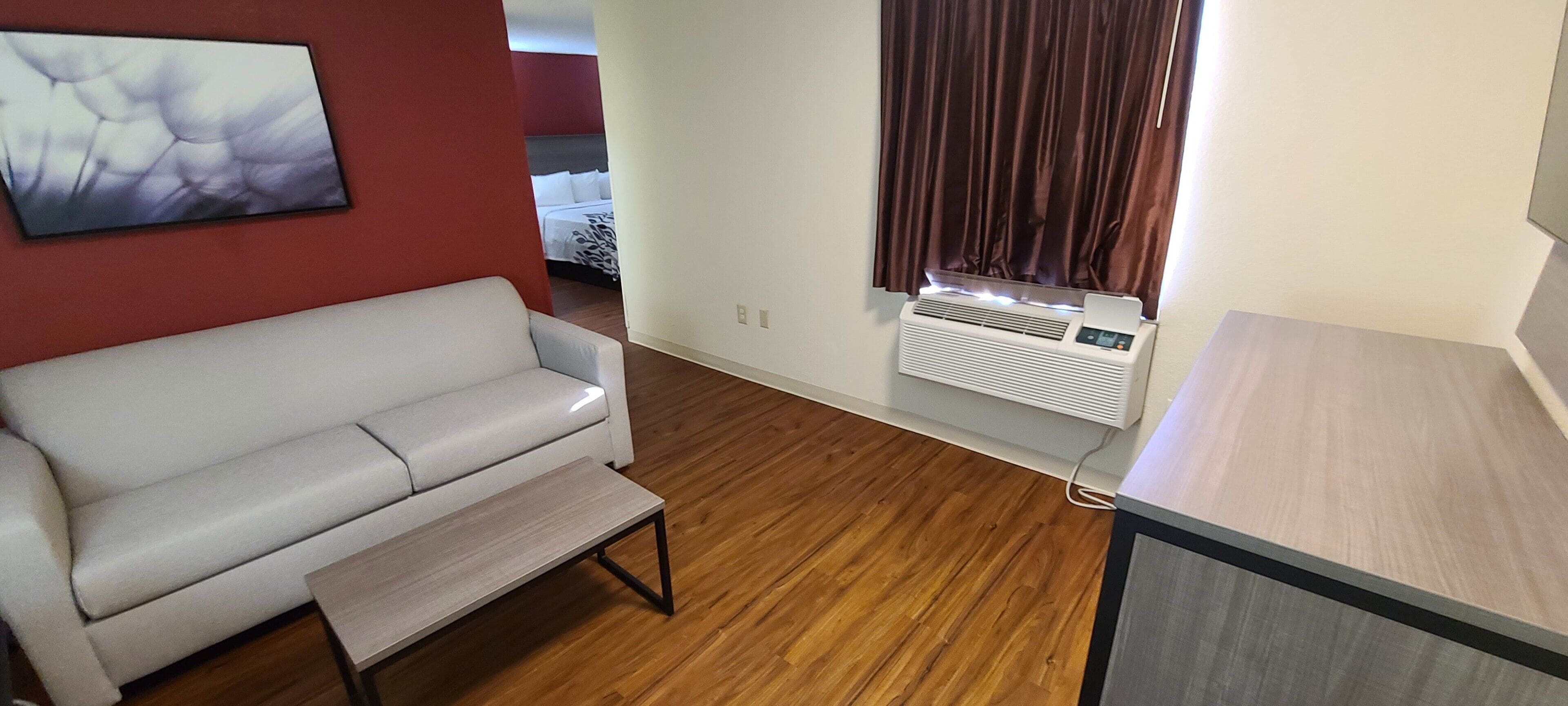 Un espacio habitable compacto con un sofá blanco, una pequeña mesa de madera, un equipo de aire acondicionado de pared y una ventana con cortinas.