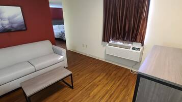 Un espacio habitable compacto con un sofá blanco, una pequeña mesa de madera, un equipo de aire acondicionado de pared y una ventana con cortinas.
