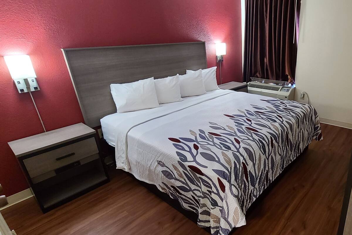 Una habitación de hotel con cama, mesitas de noche, un calentador y una pared roja.