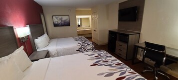 A hotel room with two beds, a desk, a chair, a TV, and a bathroom visible in the background.