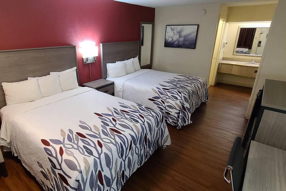 Habitación de hotel con dos camas, una silla, una mesita de noche, un espejo y un baño visible a través de una puerta abierta.