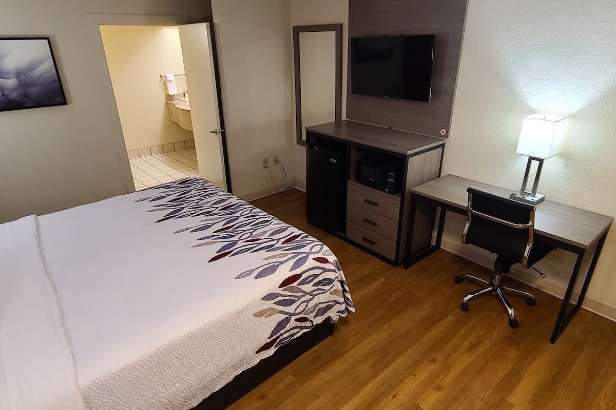 Habitación de hotel con cama, escritorio, silla, televisor y baño visible a través de una puerta abierta.