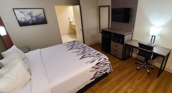 A hotel room with a bed, a desk, a chair, a TV, and a bathroom visible through an open door.
