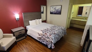 A hotel room with a bed, bedside table, chair, and a bathroom visible through an open door. - Red Roof Inn & Suites Cornelius – Lake Norman (Cornelius)