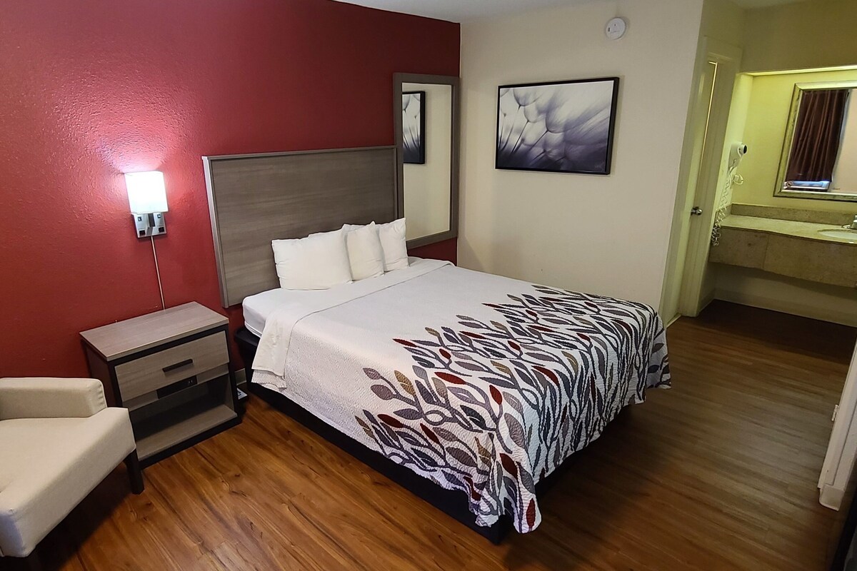Una habitación de hotel con cama, mesita de noche, silla y baño visible a través de una puerta abierta.