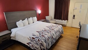 A hotel room with a large bed, a desk, a chair, a heater, and a door. - Red Roof Inn & Suites Cornelius – Lake Norman (Cornelius)