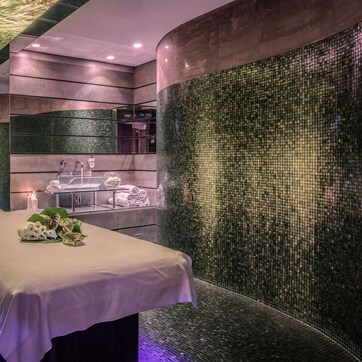 A modern spa room with a massage table, a mirror, and a tiled wall.