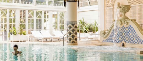 An indoor swimming pool with clear water, lounge chairs, and a decorative fountain.