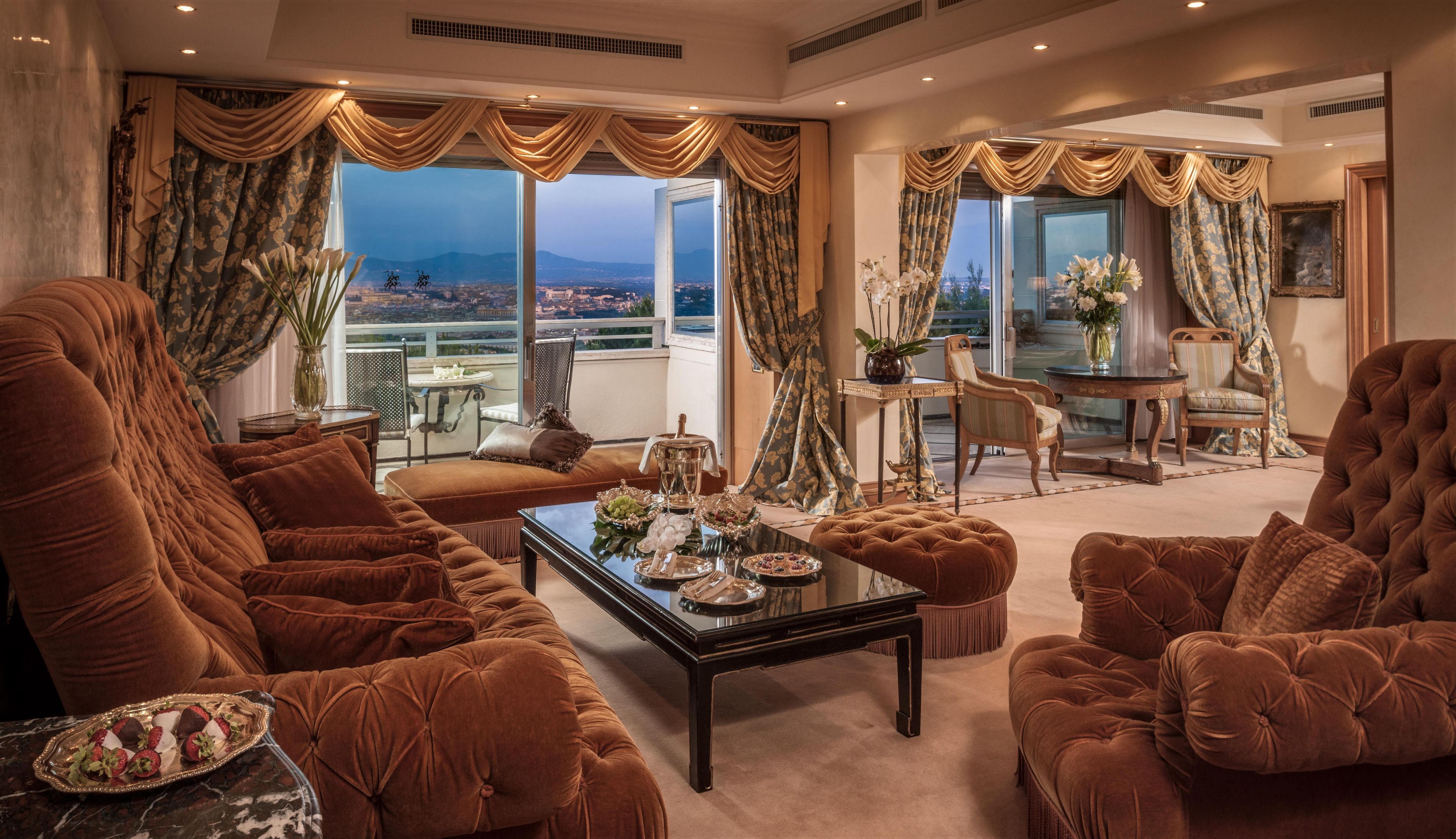 Uma sala de estar luxuosa, com sofás marrons aconcheantes, uma mesa de centro de vidro e vista para um horizonte urbano através de amplas janelas.