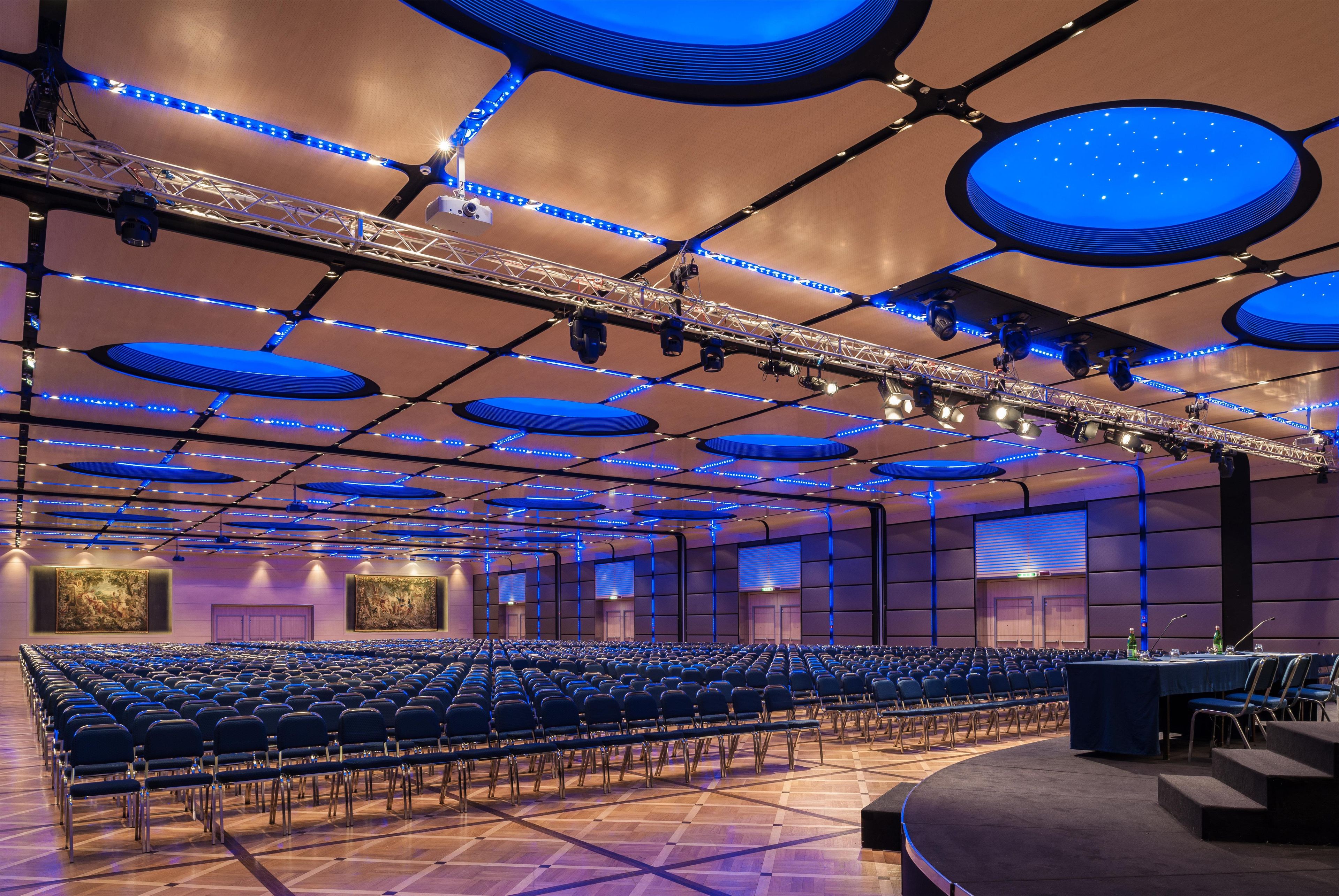 Un salón de conferencias con filas de sillas, un escenario con un atril e iluminación azul en el techo.