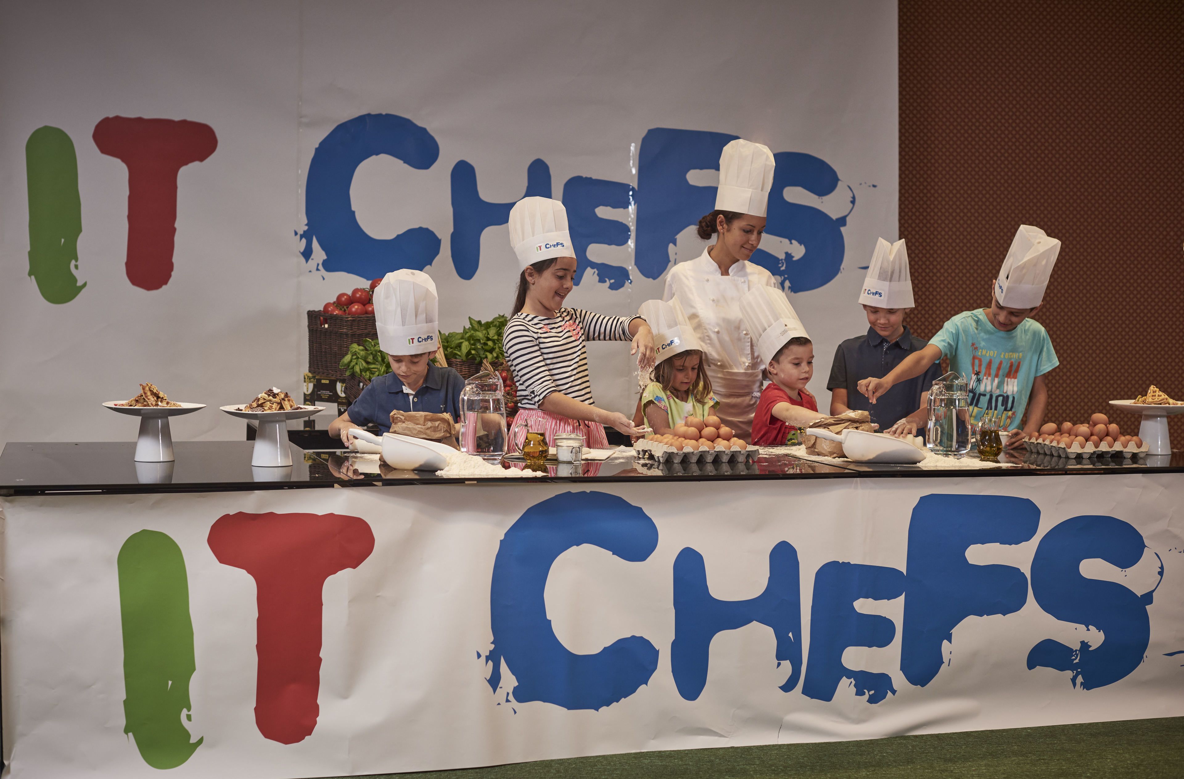 A cooking competition with participants wearing chef hats, a banner with 'IT CHEFS' text, and various cooking ingredients and utensils on the table.