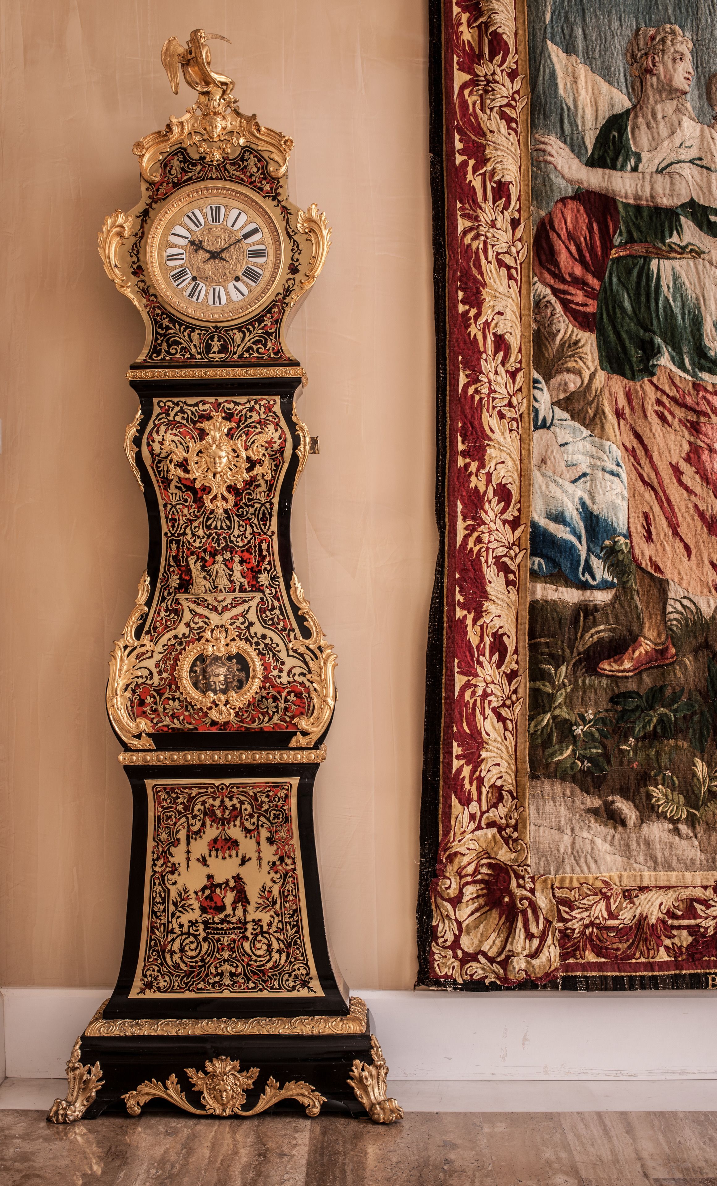 Un reloj de pie alto y ornamentado, con detalles en dorado intrincados y una esfera grande, situado frente a una pared con un cuadro de estilo clásico.