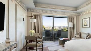 A hotel room with a large bed, a desk, a chair, a TV, a balcony with a view, and a vase with flowers.