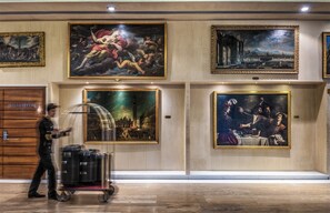 Un hall d’hôtel orné d’œuvres d’art, une porte en bois portant l’inscription « SALA TEVERE », et un chariot à bagages.