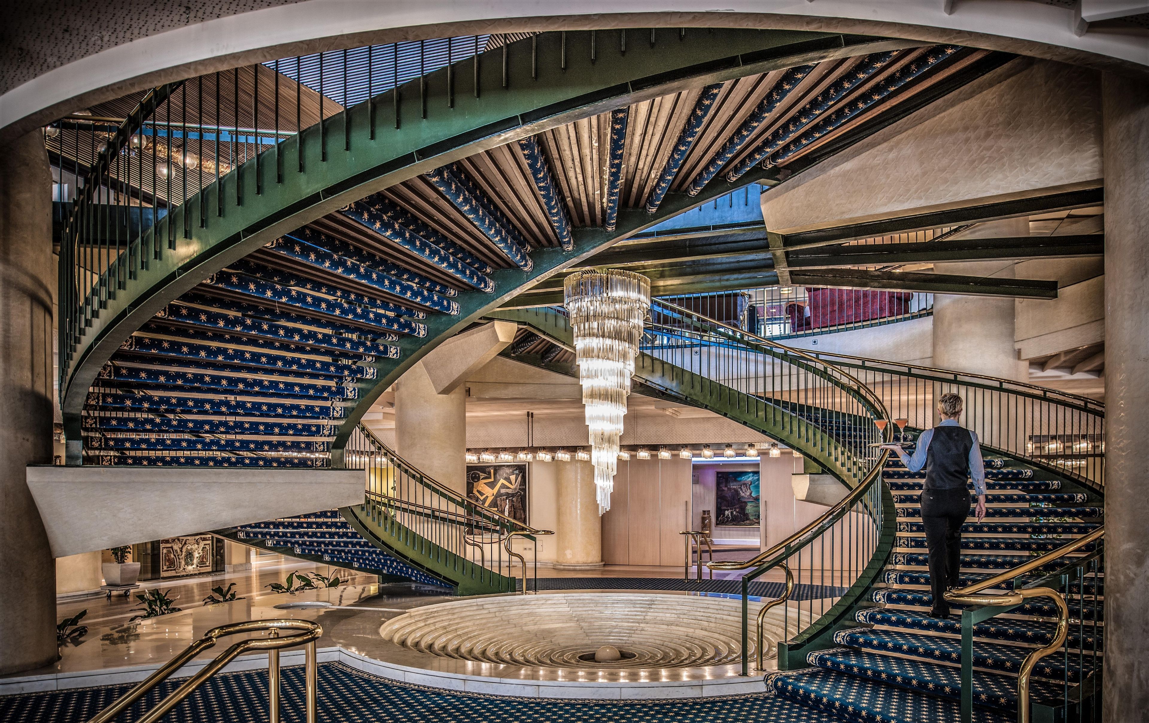 A grand spiral staircase with a chandelier, a circular fountain, and a view of the lobby area.